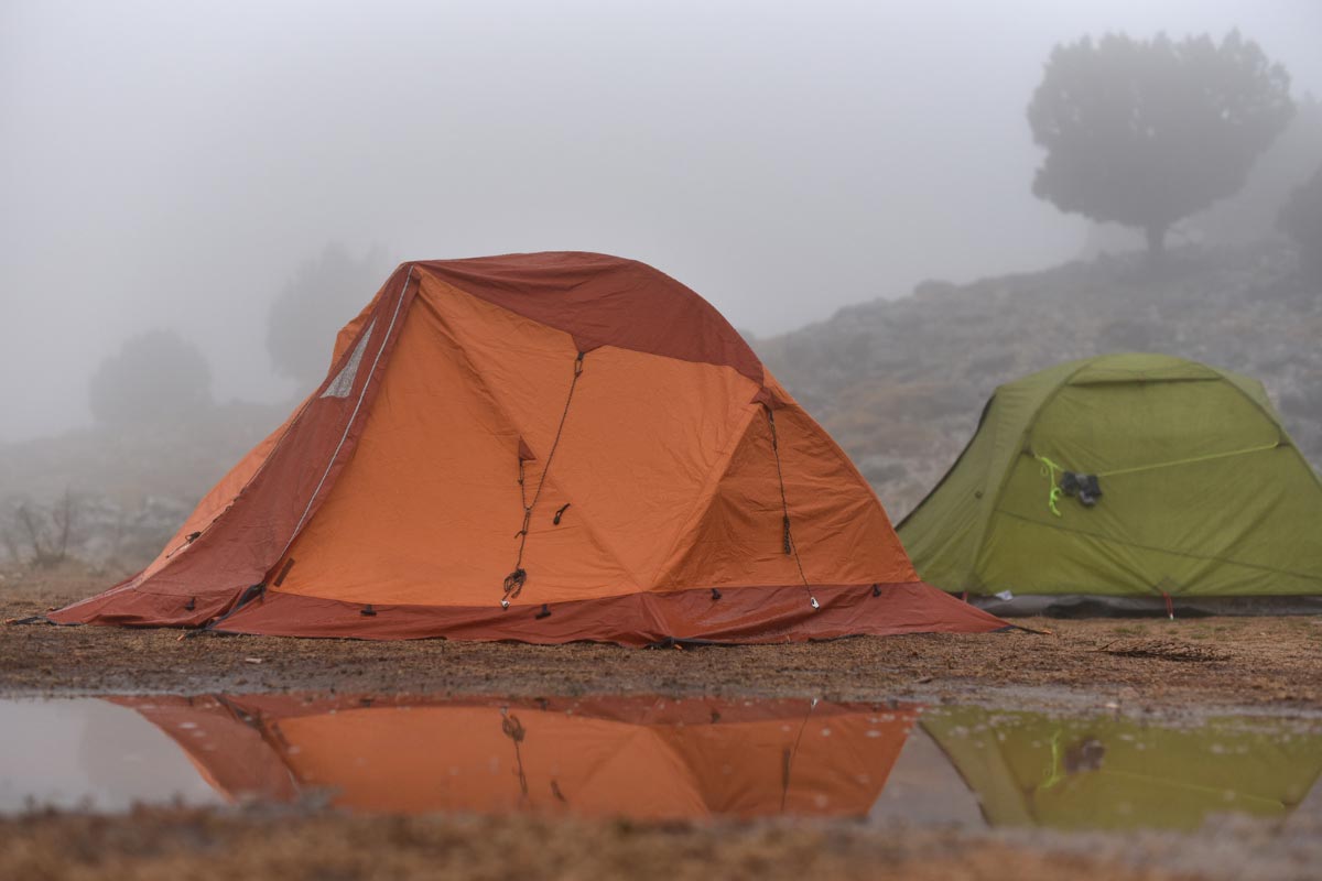 Two tents pitched near a rainy puddle with fog in the background.