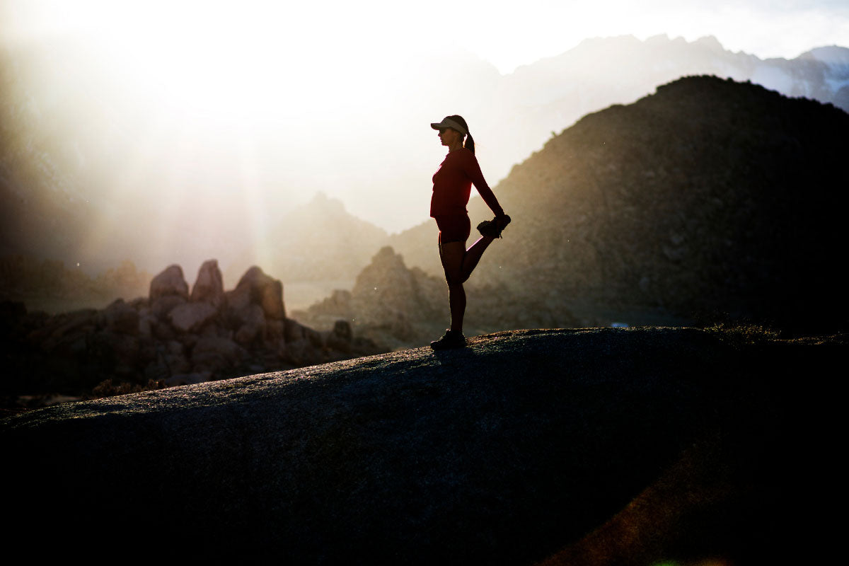 Hiker doing a thigh stretch on a mountain ridge.