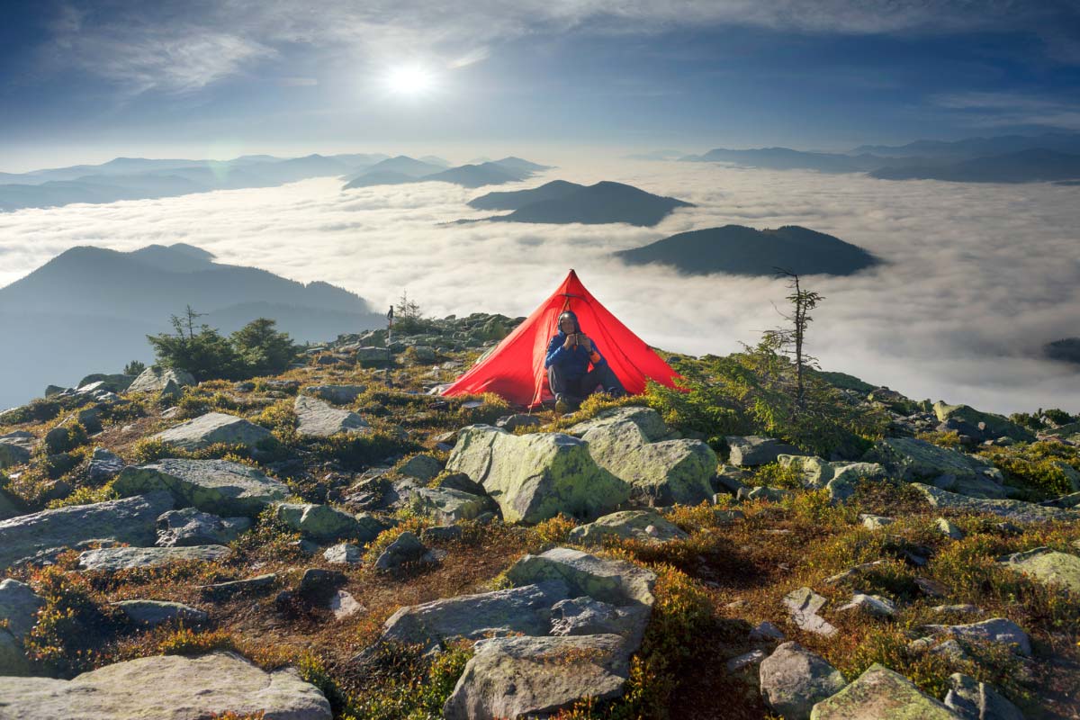 Backpacker sitting in front of an ultralight tent with mountains surrounded by clouds in the background.