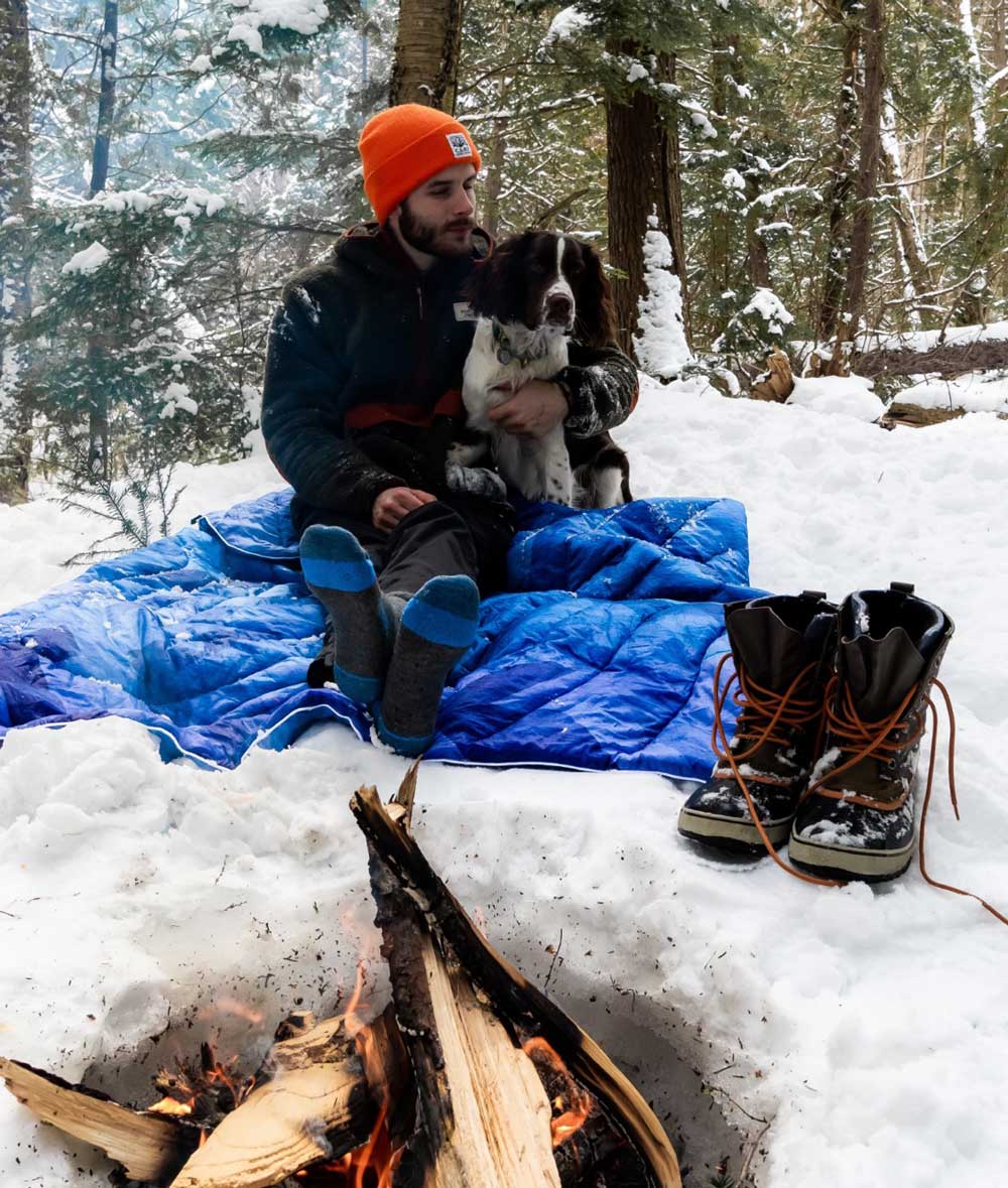 Winter camping coziness: Camper with dog by campfire, wearing Cloudline Apparel socks.