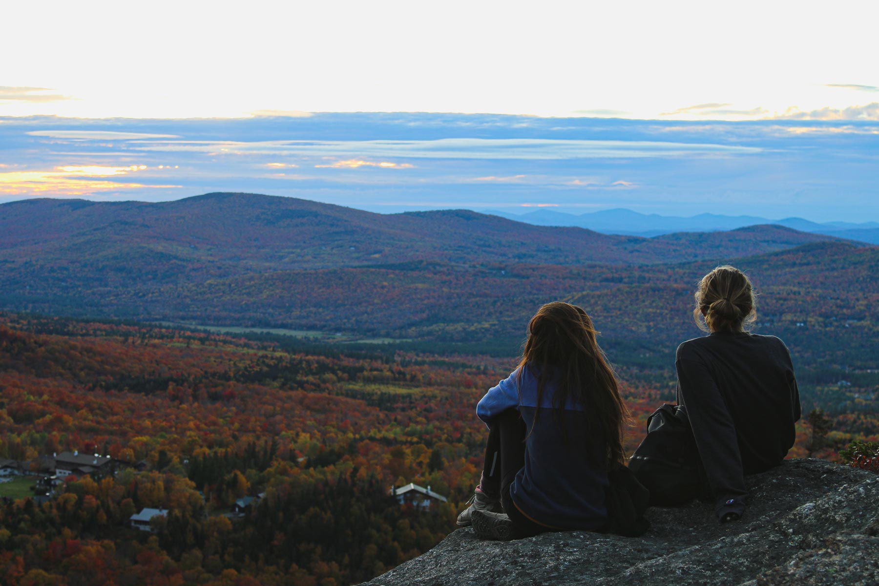 Two hikers sitting on ledge looking out over valley of fall colors.