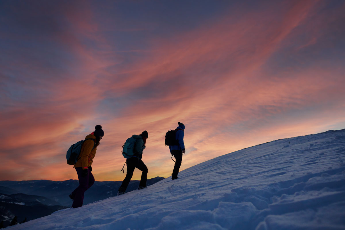 3 friends hiking up a snowy slope to see the sunset.