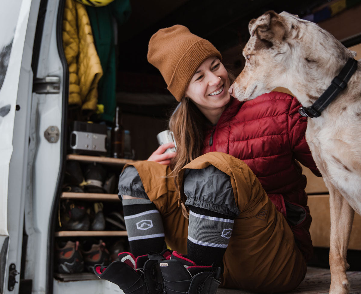 Snowboarder wearing Cloudline Apparel socks sitting in a camper van and petting a dog.
