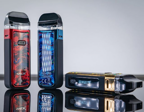 Red, blue, and gold SMOK Nord X vape devices lay atop a mirrored backdrop.