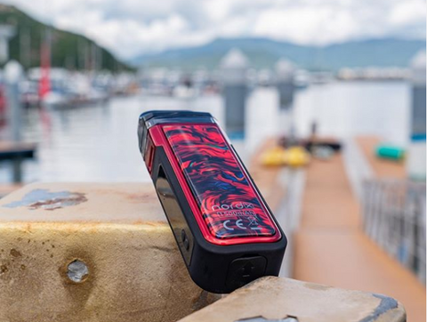 A red Nord X vape system sits on a pier with boats and water in the background.