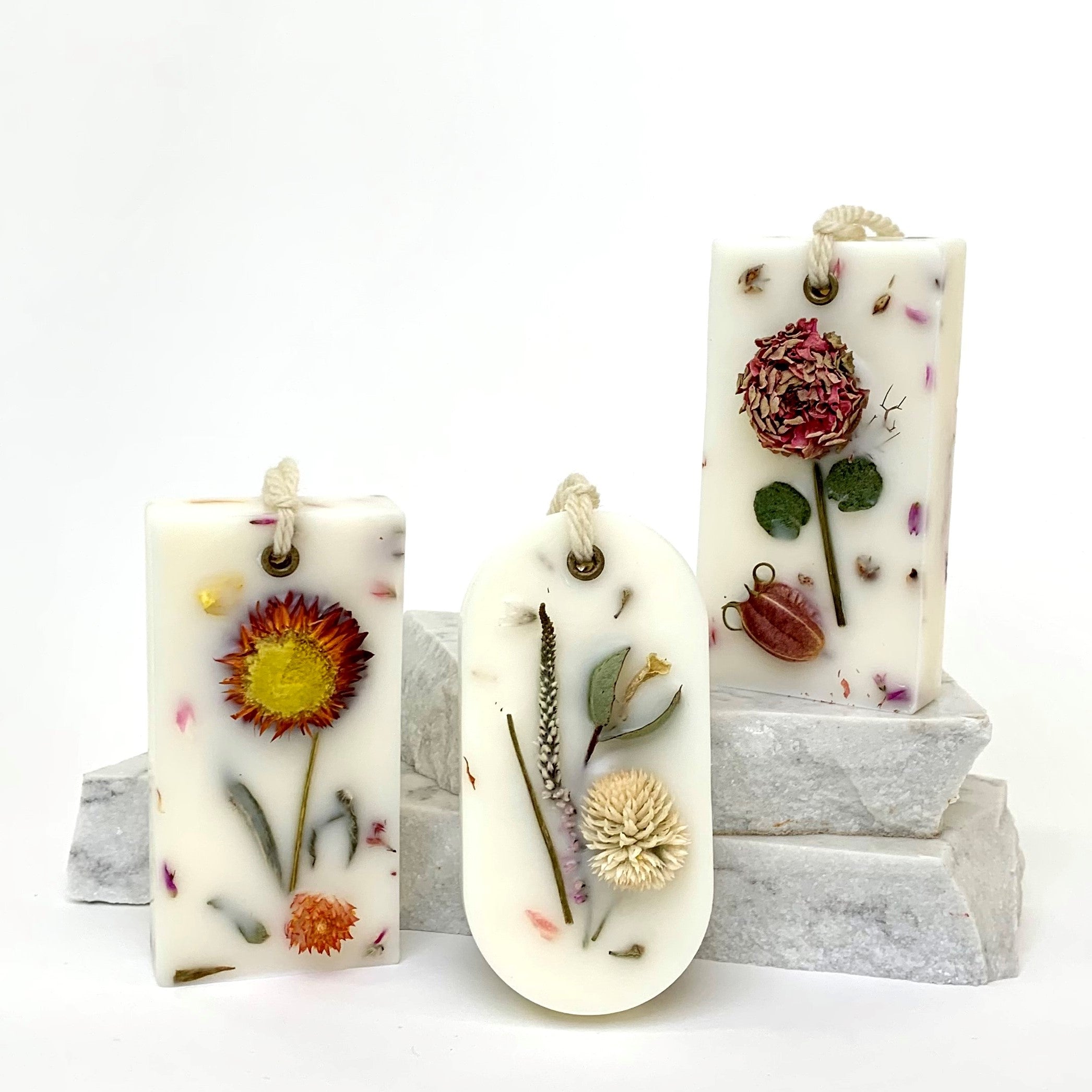 Image of 1818 Farms Botanical Wax Sachet. Three botanical waxes are pictured side by side with two in the foreground leaning on grey stones, and one in the background sitting on top of the grey stones.