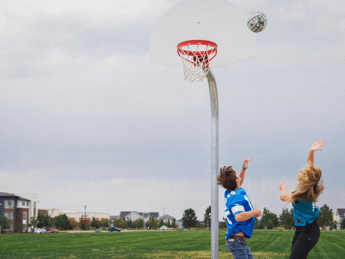 Two athletes in blue and green jerseys reach for the ball as it sails toward the basketball hoop on an outdoor court. They are captured mid-air, showcasing their competitive drive and determination, with a grassy field and houses in the distance.