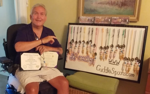 Cuddle Spoons Jewelry by Ken a Disabled person in his wheelchair holding his 2 college diplomas.