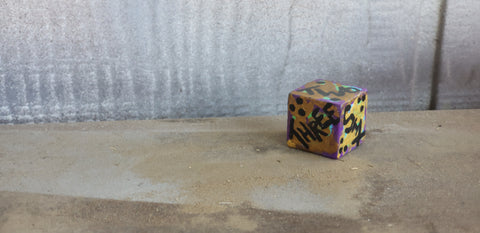 A photo of a single, painted d6 with written numbers, against a white wooden background