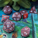 A photo of the dice set Feywild from D20Collective. The dice are made of layered multicolored wood in alternating colors of red, pink, and green. They are pictured laid across a colorful book cover