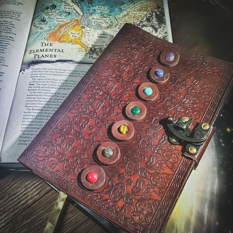 A photo of the Interplanar Travelers Leather Journal from D20Collective. The journal is a brown leather journal with tooled detailing and small stones on the cover in each color of the rainbow. A latch holds the journal closed. It is laid on a wooden table with a Dungeons and Dragons book open to a page on the Elemental Planes