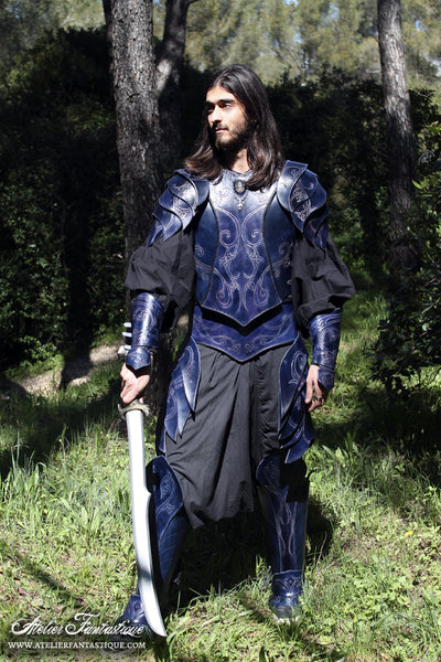 A photo of a fantasy leather armor costume. A young man with long hair wear blue leather armor, holds a sword, and looks dramatically to the side in front of a forest