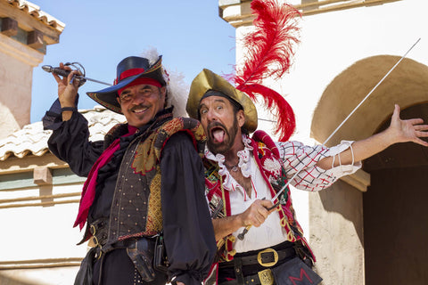 A photo of Don Juan and Miguel, comedy performers at the Arizona Renaissance Fair, dressed in Spanish Renaissance clothing, smiling at the camera and holding swords. 