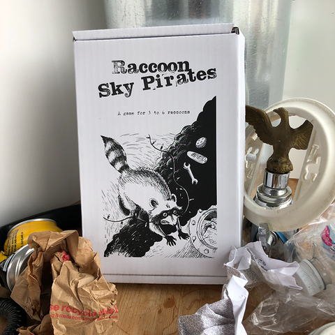 An image of the box of Raccoon Space Pirates, which has a black and white illustration of a raccoon being lowered toward the earth from a ship of some kind. The box is set on a table with various food wrappers and junk surrounding it