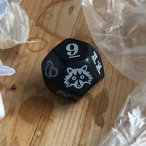 A photo of the die for Racoon Sky Pirates. It is a solid black d12 with white numbers and a racoon face in place of the numeral 12