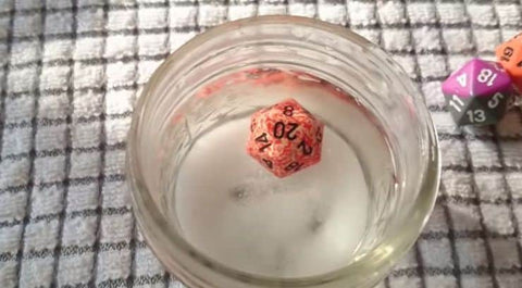 A photo of a dice in a cup of water with a lot of salt in it. The cup is set on a towel, with a couple of other dice in the background, and the die in the water is floating. 