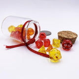 A photo of the Fire In A Bottle miniature dice set bundle from D20Collective. Several sets of semitransparent dice in yellow, orange, and red are spilling from a small glass jar with a ribbon around the neck. A cork stopper sits next to them against a white background. 