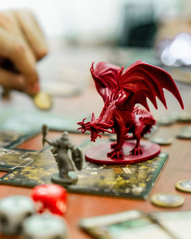 A photo of a dragon miniature placed as though in combat with several warrior miniatures, surrounded by dice and map tiles. A hand can be seen in the background.