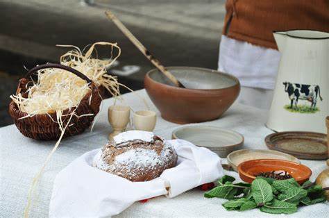 A photo of a table with a white tablecloth with rustic-looking food laid out across it. There is a loaf of bread, some spinach, a bowl and spoon, and a pitcher