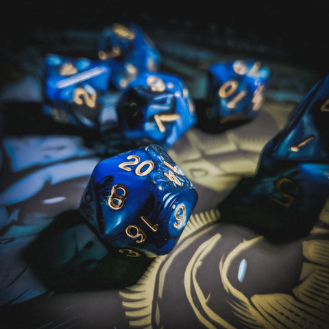 A photo of a set of dice against a dark illustrated background, of which little can be seen. The dice are blue and black with gold lettering. 