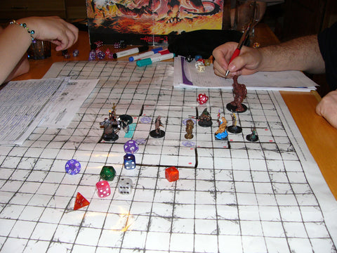 A photo of a game of Dungeons and Dragons. There is a white grid on the table with several miniatures and dice laid out on top of it, with a couple of arms and a DM screen in the background.
