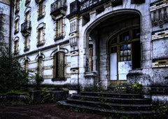 A photo of an old hotel which appears to be abandoned. There are weeds and dirt along the entrance, and a number of windows across white walls. 