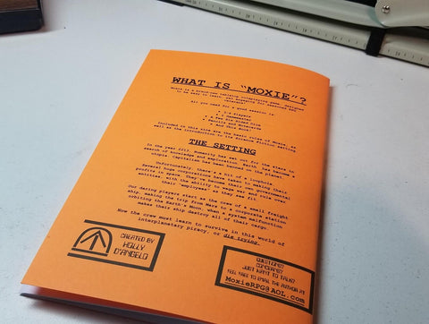 A photo of the back of the Moxie zine booklet. It s a bright orange cover with black text and no images. It sits on a white table. 