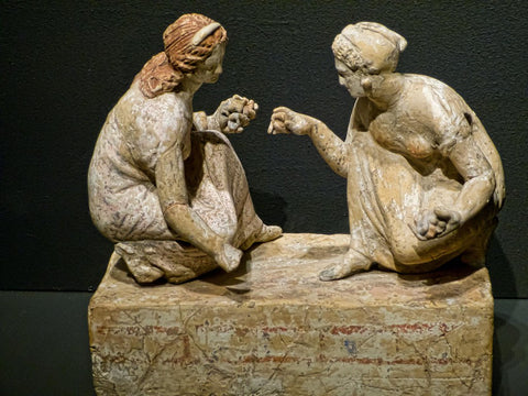 A photo of an ancient greek statue, which depicts two women kneeling, holding knuckbone dice. They are apparently playing some kind of gambling game with them. 