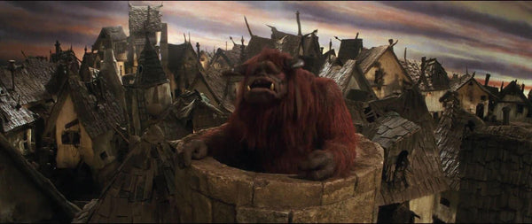 A photo of the character Ludo from the movie Labyrinth. Ludo is a large, red-furred muppet with an animal face and horns. He is popping out of a chimney overlooking a roof and the rest of a city seemingly made of rustic brown houses. The sky behind his is grey and cloudy