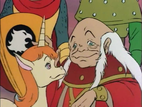A screenshot of the Dungeon Master and Uni from the DND Cartoon. it shows a short man with a bald top of his head, but long hair on the sides, in red robes. He is petting a baby unicorn with an orange mane.