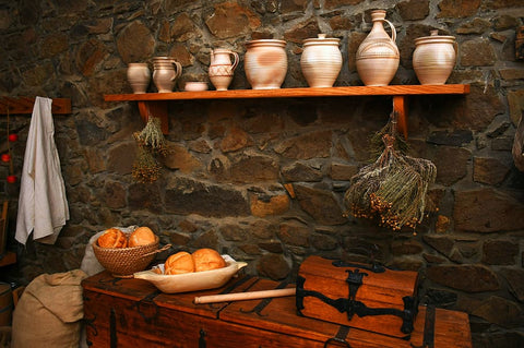 A rendering of the interior of a medieval house, with several pots on a shelf on a stone wall, above a table with food laid out
