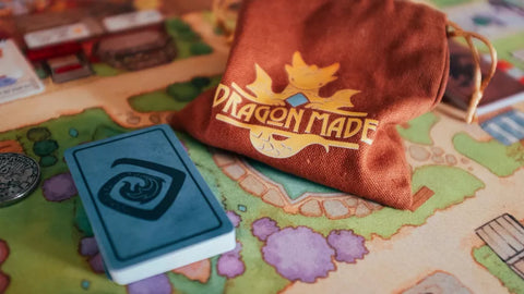 A photo of the game mat for Flamecraft, with a deck of blue cards with a swirl on them, and a small bag that says Dragonmade
