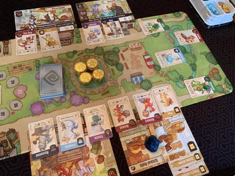 A photo of Flamecraft set up. There is a long, thin mat with an illustration of a town filled with grass, around which there are cards illustrated with cards, and various smaller dragons cards set on top of the mat