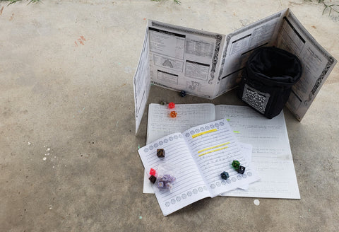 A photo of a Dungeon Masters screen, with several notebooks behind it, as well as a dice bag and some dice. The notebooks are filled with writing.