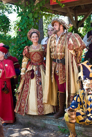 A photo of a man and woman at the texas renaissance festival. Both wear elaborate golden and white clothing and are standing on a plinth in a crowd of other people in historical garb