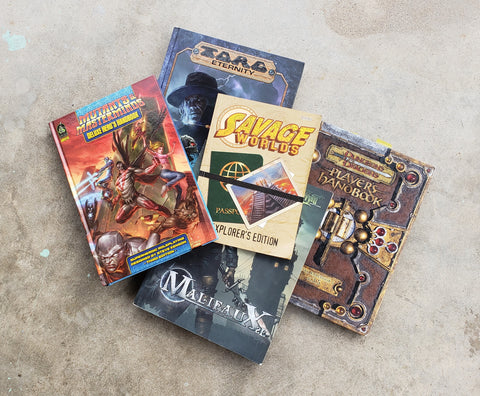 A photo of several tabletop rpgs, including Torg, Dungeons and Dragons 3.5e, Savage Worlds, Mutants and Masterminds, and Malifeaux