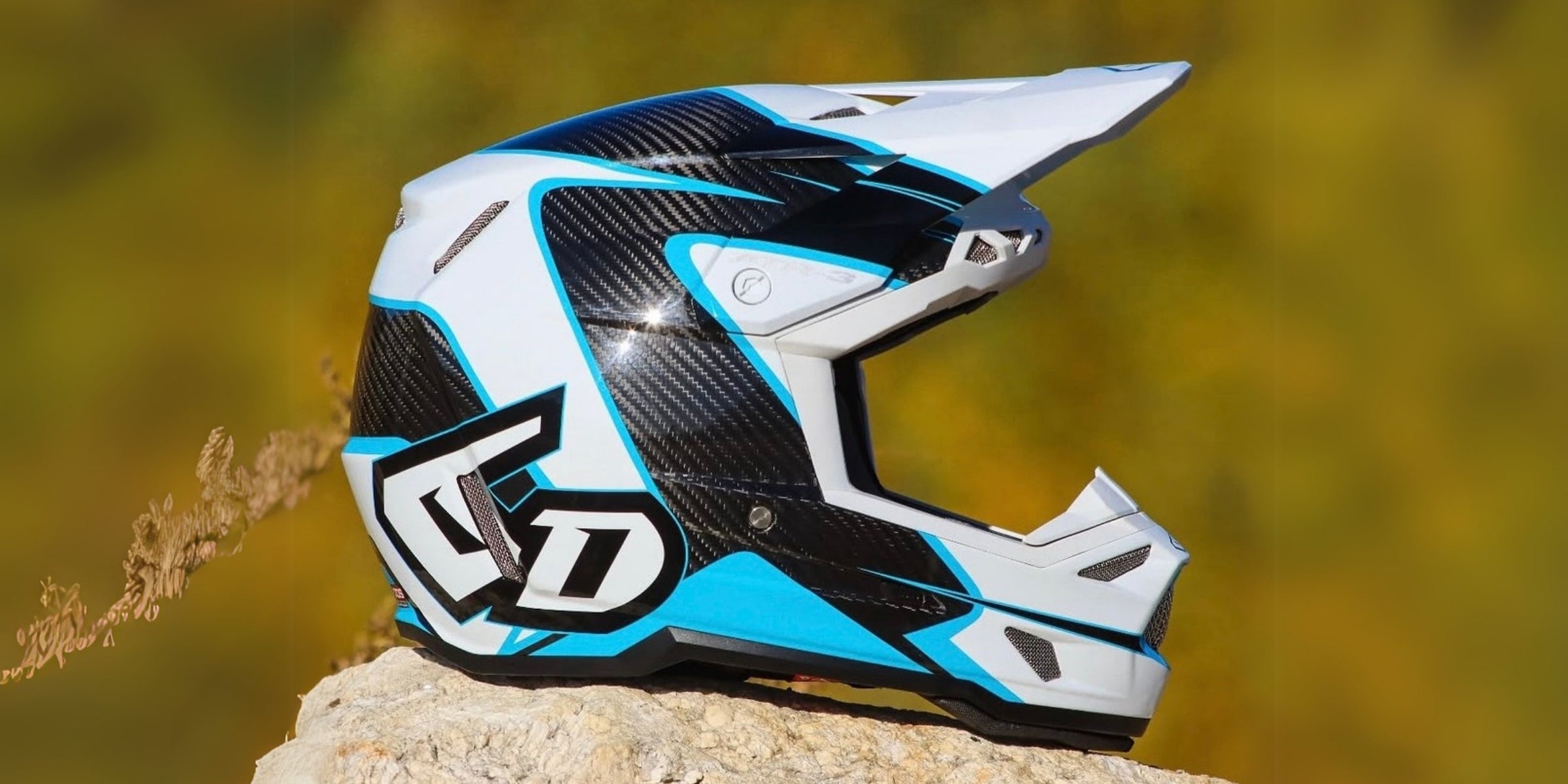 Side profile of a 6D ATR-3 motocross helmet featuring a black carbon fiber finish with white and cyan blue accents, positioned on a rock outdoors.