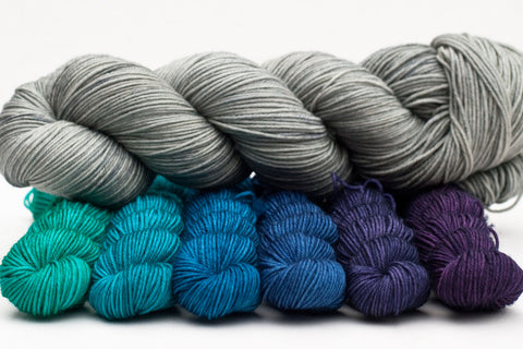 One skein of light grey yarn on top of a six mini-skein gradient in jewel tones from green through blue to purple