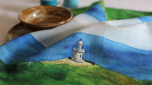 A painting on linen with a lighthouse at the end of a cliff looking into the deep blue water of the ocean.