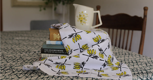 A white tea towel with yellow and black kowhai flowers in the weave like pattern. The tea towel is draped over books with a jug on the top.