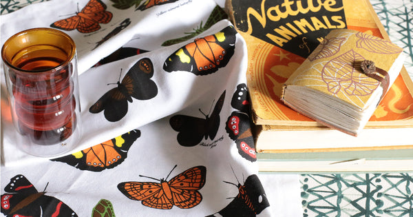 A fabric decorated with butterflies from New Zealand in oranges, bronze, brown, black, red and yellow. Lying crumpled on a table with a tea cup and books.