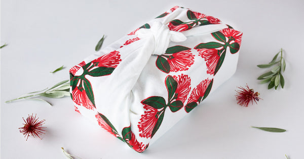 A white tea towel with a red and green print of the pohutukawa flower. The tea towel is wrapped around a present in the shape of a box.