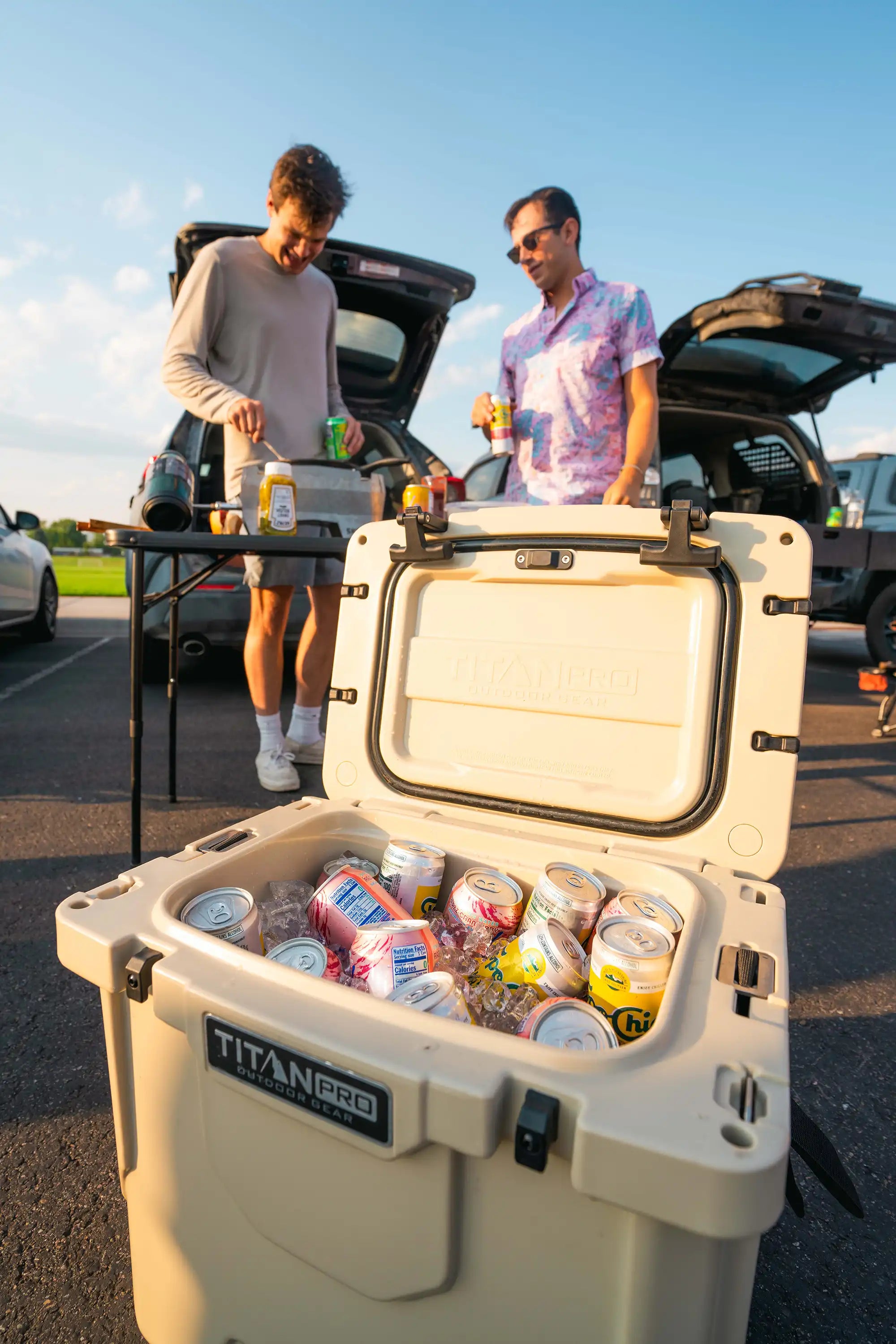 Two people grilling food at a tailgate party next to a Titan PRO Roto Hard Cooler full of drinks