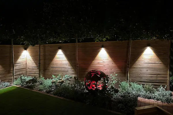 Boutique contemporary garden wth slatted wooden fencing and metal fire globe illuminated by in-lite 12 volt outdoor wall lights downlighting along the fence and planted borders.