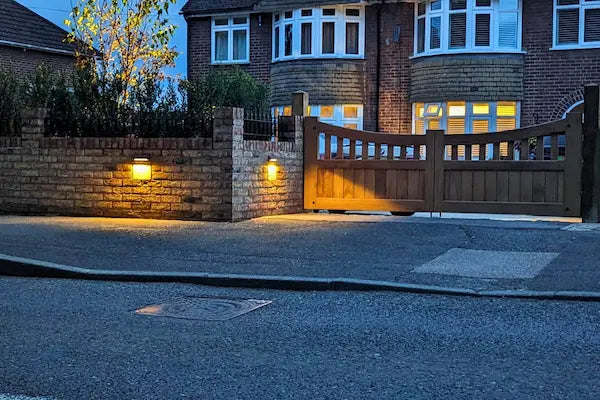 Driveway exterior lighting with in-lite 12 volt outdoor wall lights illuminating brick wall facades and highlighting iroko wooden gates to driveway entrance.