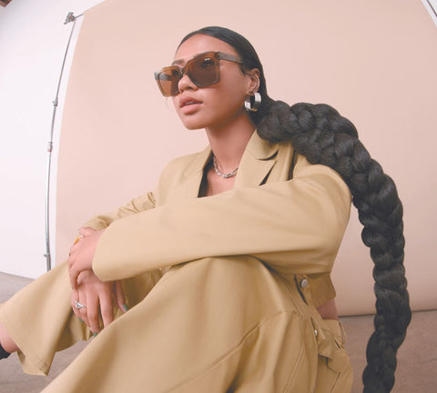 A female model sitting in front of a brown backdrop and wearing a stylish brown suit with QUAY ICY sunglasses