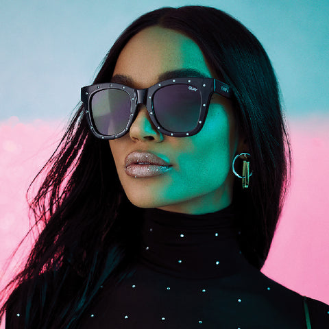 A female model posing in front of a pink and blue backdrop wearing a black outfit with QUAY AFTER HOURS black sunglasses