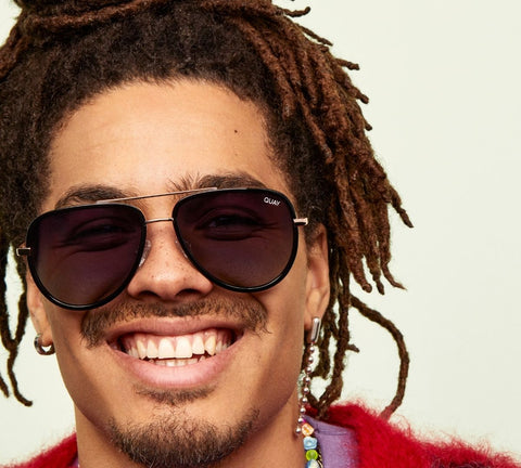 A male model smiling and wearing QUAY ALL IN sunglasses with a light olive green backdrop