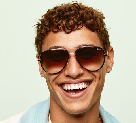 A male model with brown curly hair smiling in front of a green backdrop with QUAY ALL IN tortoise sunglasses