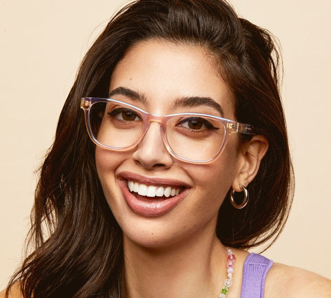 A female model smiling in front of a brown backdrop wearing QUAY HARDWIRE MINI clear glasses